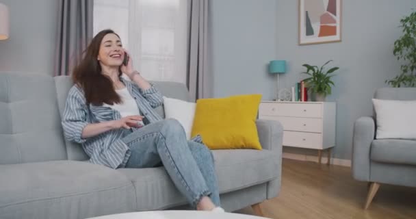 Vacker vit glad ung kvinna sitter ner på soffan i mysiga vardagsrum och chatta på smartphone. Glädjande söt flicka ler och talar på mobilen hemma. Fritidsidé — Stockvideo