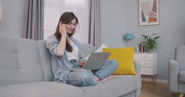 Glada vackra kaukasiska mörkhåriga flicka som arbetar på laptop inomhus hemma. Ganska glad ung kvinna skriva och bläddra på datorn medan du sitter på soffan i mysigt vardagsrum. Frilansbegreppet — Stockvideo