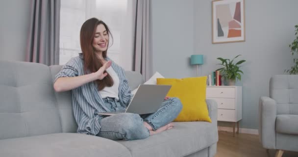 Весела гарна дівчина, яка розмовляє на ноутбуці в приміщенні. Радісна красива кавказька молода жінка махає рукою і розмовляє через комп'ютерну веб-камеру, сидячи вдома на дивані. Концепція спокою — стокове відео