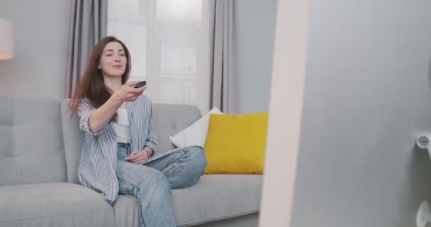 Hezká kavkazská šťastná mladá žena sedící v obývacím pokoji na pohovce a zapínající televizi. Krásná dívka v dobré náladě přepínání kanálů s dálkovým ovládáním při sledování televize doma. Koncept odpočinku — Stock video