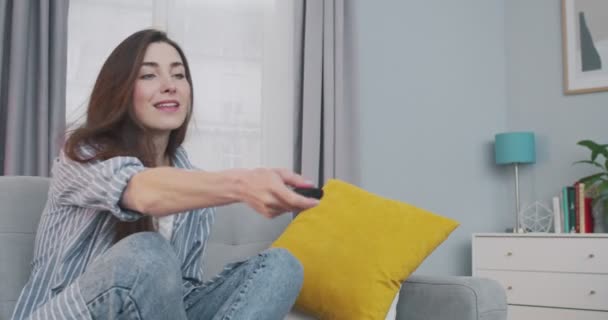 Jolie jeune femme heureuse caucasienne assise sur le canapé dans le salon et allume la télévision. Portrait de belle fille de bonne humeur reposant à la maison seul tout en regardant la télévision. Concept de loisirs — Video