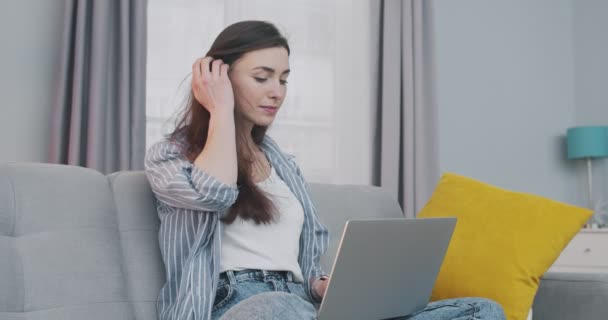 Concentrada bela menina de cabelos escuros caucasiana trabalhando em laptop dentro de casa. Mulher muito jovem digitando e tocando no computador enquanto sentado no sofá cinza na acolhedora sala de estar. Conceito freelance — Vídeo de Stock