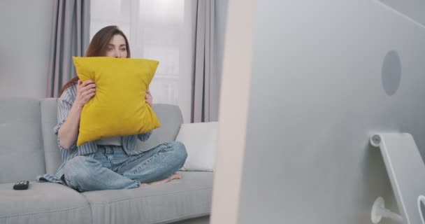 Schöne verängstigte Mädchen, die allein zu Hause ausruhen und fernsehen. Hübsche kaukasische verängstigte junge Frau sitzt auf grauem Sofa im Wohnzimmer und schaut Horrorfilm im Fernsehen. Freizeitkonzept — Stockvideo