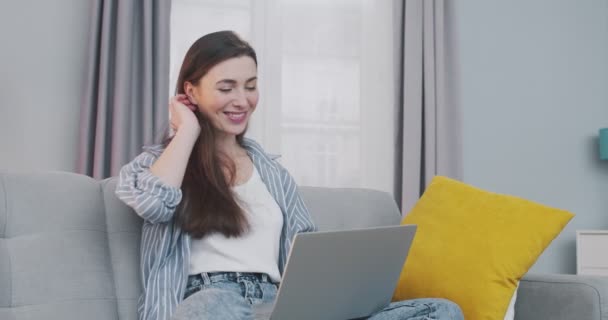 Vreugdevolle mooie blanke jonge vrouw glimlachen en spreken via computer webcam terwijl zitten op de bank in de kamer thuis. Gelukkig mooi meisje met een video gesprek op de laptop. Vrijetijdsconcept — Stockvideo