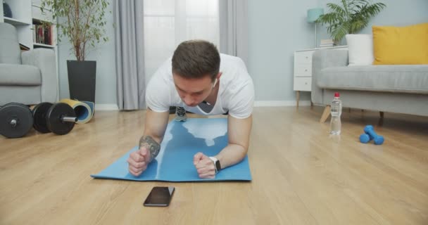 Entrenamiento de tipo deportivo, haciendo ejercicio mientras escucha música en los auriculares en casa. Deportista concentrado haciendo un ejercicio de tablón en la estera utilizando una aplicación smartwatch. Salud, deportes, concepto de bienestar. — Vídeo de stock