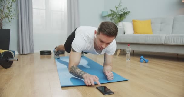 Muskulös kille gör fitness och träna på en matta, lyssnar på musik i trådlösa hörlurar. Idrottare med mycket uthållighet gör en planka motion med hjälp av en smartphone app, timing i ett stoppur. — Stockvideo