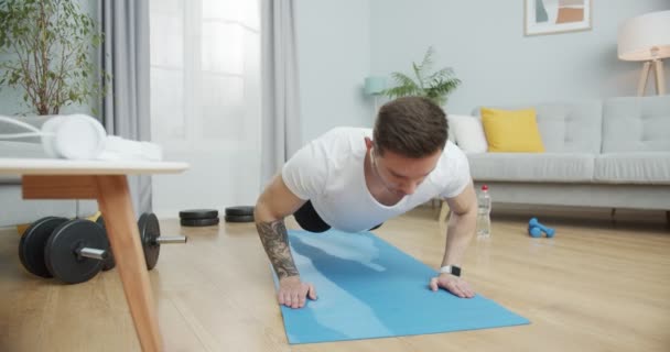 한 젊은 코카서스 운동 선수가 무선 이어폰으로 음악을 듣기 위해 터벅터벅 뛰고 있습니다. 강력 한 운동 선수인 그는 집에서 운동을 하고, 훈련을 받고, 건강을 유지 한다. 건강하고 부유 한 개념. — 비디오