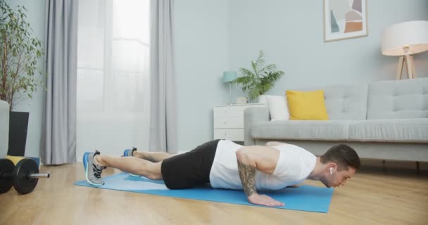 Un homme fort faisant des exercices d'entraînement à la maison. Un sportif qui fait des pompes, des pompes, écoute de la musique dans les écouteurs dans le salon avec un intérieur minimaliste. Un concept de formation et d'entraînement. — Video