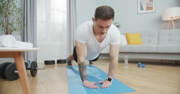 Un homme athlétique faisant de l'exercice et de remise en forme, écoutant de la musique dans les écouteurs à la maison. Un mâle musclé faisant des pompes triangulaires sur un tapis. Un concept sportif et d'entraînement d'un mode de vie sain. — Video
