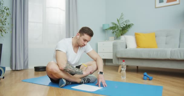 Un deportista concentrado tomando notas en un papel, registrando los resultados y los cambios. Un joven guapo contando y anotando calorías, haciendo un horario. Un concepto de entrenamiento y entrenamiento. — Vídeos de Stock