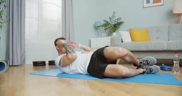 Lichaamstraining binnenshuis tijdens pandemie, isolatieperiode, quarantaine. Jongeman doet sit-ups, tilt hoofd en schouders van de vloer, draait op de knieën. Sport, fitness, trainingsconcept. — Stockvideo