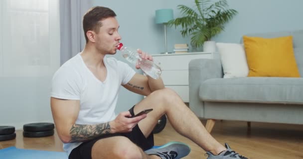 Spocony, zmęczony sportowiec pijący butelkę wody po treningu, fitness, gimnastyka, aerobik. Muskularny mężczyzna korzystający z telefonu, surfujący po mediach społecznościowych, sprawdzający wiadomości, siedzący na podłodze. Uprawiać sport w domu. — Wideo stockowe