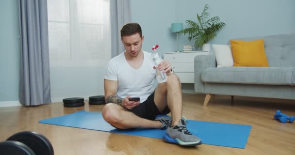Stark idrottsman dricka flaska vatten efter träning hemma. Attraktiv kille som använder smartphone, bläddrar genom Internet, sociala nätverk, media, sitiering på golvet i vardagsrummet. En hälsosam livsstil. — Stockvideo
