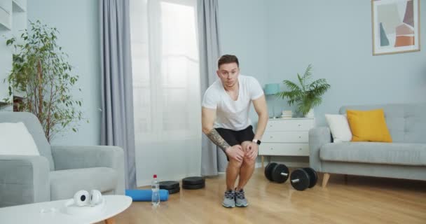 Een fitnesstrainer in sportkleding trainen, rekken om fit te blijven thuis. Een aantrekkelijke man die kniekringoefeningen doet, die voor de training opduikt. Welzijn, gezondheidsconcept. — Stockvideo