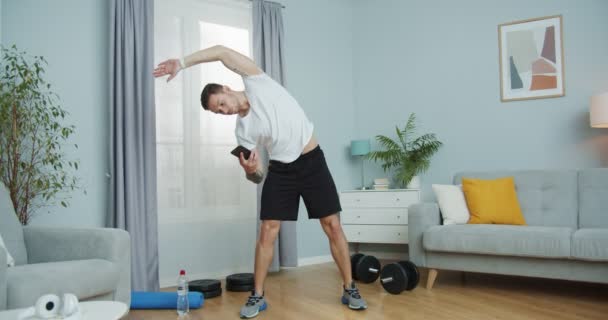 Un tipo caucásico atractivo haciendo aeróbic, gimnasia en casa. Un joven musculoso calentándose, estirándose, haciendo ejercicios de curvas de pie disfrutando del tiempo libre. Bienestar y concepto sanitario. — Vídeos de Stock