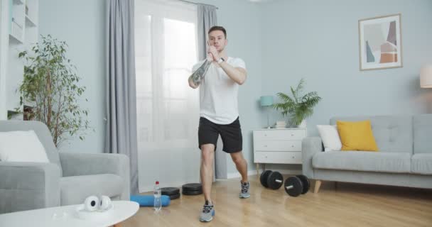 Un hombre caucásico desgarrado haciendo ejercicio, atletismo en casa. Un hombre fuerte cojeando hacia arriba, haciendo repeticiones de ejercicios de embestidas para crecer. Perder libras, perfilarse, mantenerse en forma, seguir un estilo de vida saludable — Vídeos de Stock