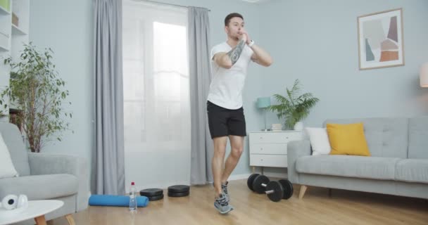 Un joven instructor de fitness entrenando, ejercitándose en casa. Un macho musculoso deportivo realizando ejercicio en la parte inferior del cuerpo, saltando saltos, sentadillas pop en la habitación. Automotivación, salud, concepto deportivo. — Vídeos de Stock