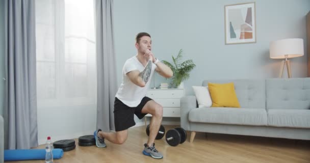 若いフィットネストレーナーは、家庭で働く、暖まるストレッチ。下半身運動をするスポーティーな筋肉の男は、リビングルームで肺をジャンプします。自己動機、医療、スポーツの概念. — ストック動画