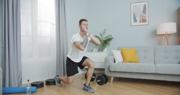 Fiatal fitnesz edző nyújtózkodik, bemelegít, otthon edz. Egy sportos, izmos fickó, aki alsó testmozgást végez, ugró tüdővel a nappaliban. Önmotiváció, egészségügy, sport koncepció. — Stock videók