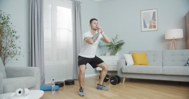 Un joven deportista haciendo ejercicios de muslos en cuclillas en casa. Un fuerte entrenamiento masculino para tonificar los glúteos, fortalecer los músculos de las piernas en la sala de estar. Salud, deportes y un concepto de estilo de vida rico. — Vídeos de Stock