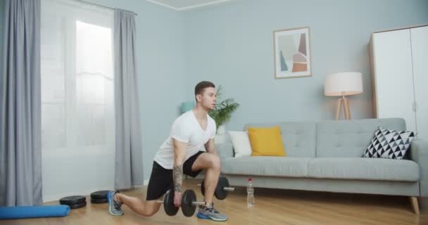 Genç fitness antrenörü ısınıyor, evde çalışıyor. Yakışıklı sporcu alt vücut egzersizi yapıyor, oturma odasında dambıllarla saldırıyor. Motivasyon, sağlık, spor konsepti. — Stok video