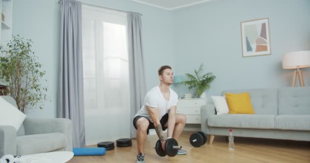 Deportivo tipo caucásico haciendo ejercicio en casa durante la pandemia. Hombre atlético en forma haciendo ejercicios en cuclillas con una mancuerna para fortalecer los músculos, tonificar, en forma. Entrenamiento en interiores. Autoaislamiento, concepto de salud. — Vídeo de stock