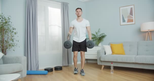 Mladý fitness instruktor dělá fitness, cvičí doma. Pohledný sportovec, který cvičí v dolní části těla, opakuje výpad s činkami v obýváku. Motivace, zdravotní péče, sportovní koncept. — Stock video