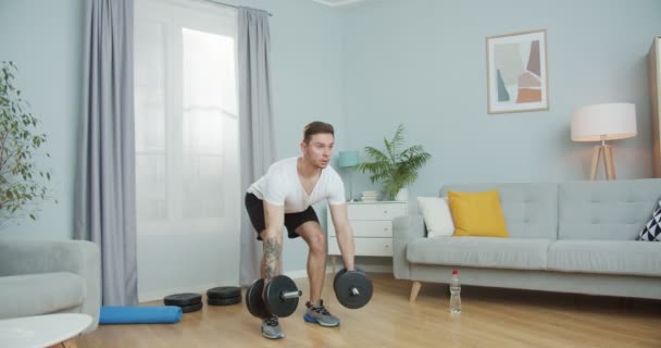 Mladý atletický chlapík dělá doma sportovní fitness. Svalnatý, svalnatý muž provádějící ohýbání, cvičení Barbell Row s činkami, činky na biceps. Vnitřní cvičení, atletika během karantény. — Stock video