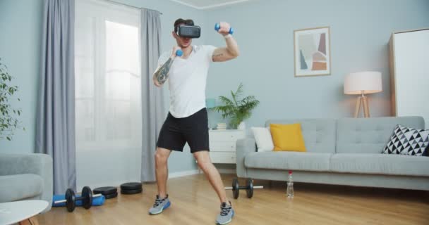 VR gözlüklü beyaz tenli antrenör evde çalışıyor. Çekici, formda bir adam boksu siber uzayda ağırlık kaldırarak dövüşüyor. Sanal gerçeklik oyunları. Dijital fütüristik spor, siber uzay kavramı. — Stok video