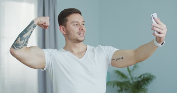 Joven chico fuerte tomando foto selfie para las redes sociales después de entrenar en casa. Sonriente deportista atractivo mostrando bíceps, posando en la cámara del teléfono inteligente. Selfie, foto, concepto de redes sociales — Vídeo de stock