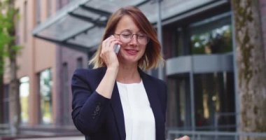 Modern ofis binasının yakınında resmi giysiler içinde yürüyen beyaz bir kadının portresi. Gözlüklü, gülümseyen bir iş kadını meslektaşıyla konuşuyor, etrafa bakıyor. Çalışma, iletişim konsepti.
