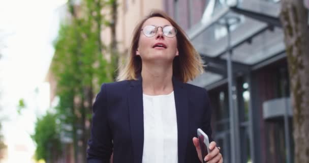 Urocza, biała bizneswoman w okularach, spacerująca w pobliżu nowoczesnego biurowca. Dorosła pracownica w formalnych ciuchach za pomocą smartfona do pisania SMS-ów, czatu w Internecie. Media społecznościowe, koncepcja technologii. — Wideo stockowe