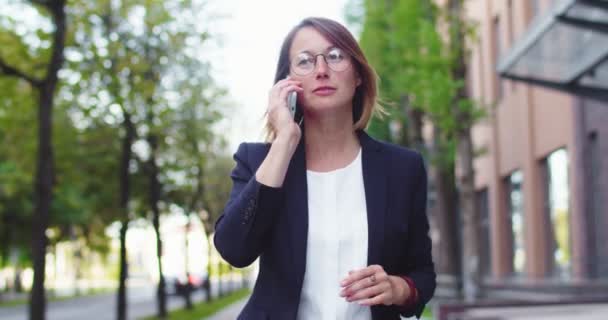 Okouzlující dospělá žena ve společenském obleku procházející se v blízkosti moderní kancelářské budovy v obchodní čtvrti. Usmívající se zaměstnavatelka mluví s partnerkou po telefonu, spravuje vlasy. Technologie, přístroje, komunikace. — Stock video
