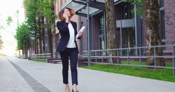 Úspěšná dospělá podnikatelka kráčející v blízkosti moderní kancelářské budovy, držící dokumenty a notebook. Pěkně vypadající běloška v obleku, jak telefonuje s klientkou. Práce, profese. — Stock video