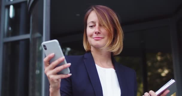 ภาพของผู้หญิงผู้ใหญ่ที่ดูดีในชุดสูทอย่างเป็นทางการยืนอยู่ใกล้กับอาคารสํานักงานที่ทันสมัย ยิ้มสาวธุรกิจคนขาวใช้สมาร์ทโฟนเลื่อนฟีดข่าว สื่อสังคม อินเทอร์เน็ต เทคโนโลยี . — วีดีโอสต็อก