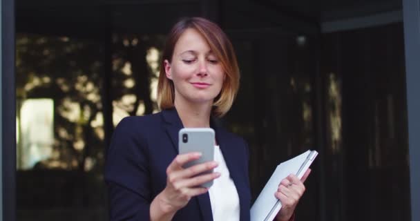 Attractive Caucasian businesswoman standing outside modern corporation building and using smartphone. Cheerful adult female boss chatting, texting with friend and smiling. Social media, Internet. — Stock Video