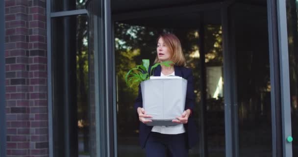 Разочарованная взрослая женщина уволена, выходит из офисного здания и смотрит в камеру. Уволенная белая женщина в официальном костюме держит картонную коробку с рабочими вещами, растение. Работа, увольнение. — стоковое видео