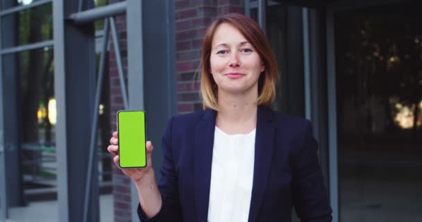 Hezká podnikatelka stojící před moderní kancelářskou budovou. Dospělý běloška žena šéf drží smartphone se zelenou obrazovkou venku. Chroma klíč, miniaplikace, koncept technologie. — Stock video