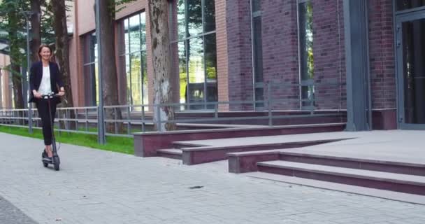 Attrayant adulte femme d'affaires en costume formel s'amuser à l'extérieur de l'immeuble de bureaux moderne. Joyeuse ouvrière caucasienne chevauchant scooter électrique coup de pied et souriant. Technologie, concept d'écologie. — Video