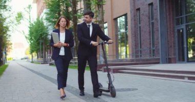 Modern ofis binalarının yanında yürüyen kadın ve erkek meslektaşlar. Elektrikli scooter kullanan güzel bir işadamı ve elinde dizüstü bilgisayar olan resmi giysili bir kadın. Teknoloji konsepti.