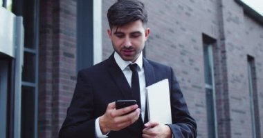 Modern şirket binasının önünde duran kendinden emin beyaz erkek işçi. Akıllı genç iş adamı dizüstü bilgisayar tutuyor ve akıllı bir şekilde mesajlaşıyor, sohbet ediyor. Teknoloji, iş konsepti.