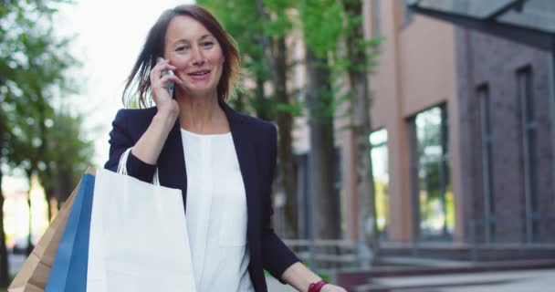 Ganska vit kvinna i formell kostym går med inköp i paket. Leende affärsdam som ringer till college, pratar och skrattar. Teknik, shopping, jobb, hobby. Bakgrund till kontorsbyggnaden. — Stockvideo