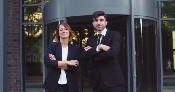 Un homme et une femme prospères debout à l'extérieur d'un immeuble moderne. Collèges masculins et féminins élégants en costumes formels croisant les bras et regardant la caméra. Entreprise, concept de partenariat. — Video