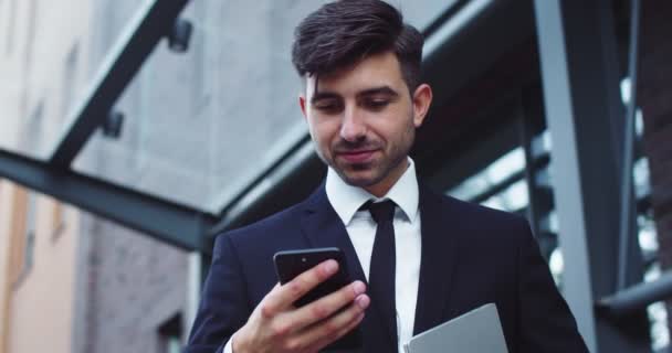Un jeune homme d'affaires confiant utilisant un smartphone à l'extérieur d'un immeuble de bureaux moderne. Beau patron masculin en costume formel défilement flux de nouvelles, messages texte. Technologies, concept de gadgets. — Video