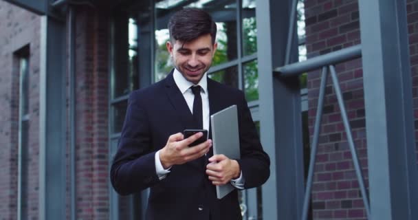 Charmig vit affärsman i formell kostym utanför modern kontorsbyggnad. Unga manliga entreprenör bläddra nyhetsflöde, läsa meddelanden på digital smartphone. Teknik, entreprenörskap. — Stockvideo