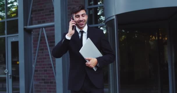 Веселый белый предприниматель с ноутбуком, выходящим из современного здания корпорации. Молодой бизнесмен разговаривает с другом, коллегой по телефону. Профессия, концепция предпринимательства. — стоковое видео