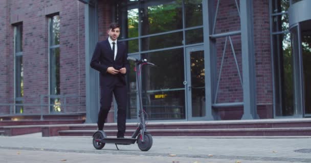 Homme d'affaires caucasien élégant venant à scooter électrique kick près de l'immeuble de l'entreprise moderne. Jeune employé masculin attrayant utilisant un smartphone pour commencer à monter. Technologie, concept d'écologie. — Video