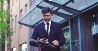 Scooter 'ı çalıştırmak için akıllı telefon uygulaması kullanan yakışıklı beyaz erkek yöneticinin portresi. Genç iş adamı dışarıda ekolojik nakliye yapıyor. Ekoloji, iş, ulaşım kavramı.