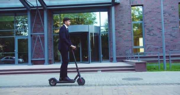 現代のオフィスビルの近くに電動キックスクーターに乗って良い見て白人実業家。成功した大人の男性起業家は、生態系の輸送によって動作するようになる。よく、成功、仕事. — ストック動画