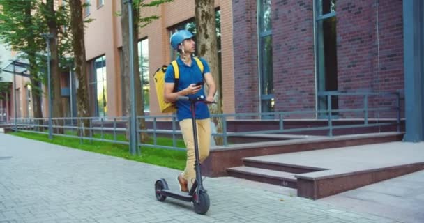 Yakışıklı beyaz erkek kurye scooter 'la gidiyor ve müşteri adresini arıyor. Kasklı genç teslimatçı teslimat yerini bulmak için akıllı telefonu kullanıyor. Meslek, teknoloji, uygulama kavramı. — Stok video